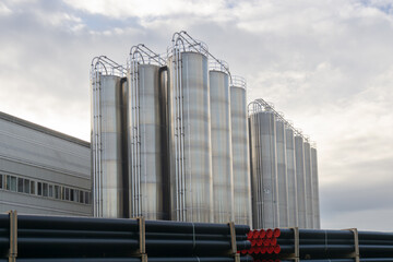 Large stainless steel industrial silos at a factory site with stacked plastic pipes in the...
