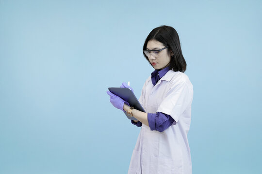 Focused Asian female scientist or doctor wearlab coat, writing data on a digital tablet, symbolizing meticulous research and modern record-keeping in blue studio background