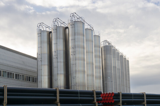 Large stainless steel industrial silos at a factory site with stacked plastic pipes in the foreground, representing manufacturing and industrial infrastructure.