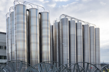 Large group of industrial stainless steel silos used for storage in a factory or processing facility, with ladders and safety railings visible on top