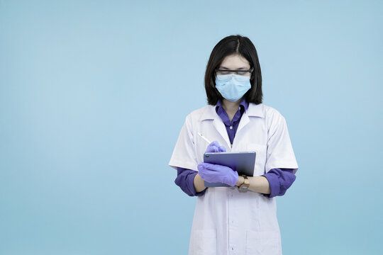 Focused Asian female scientist or doctor wearlab coat, writing data on a digital tablet, symbolizing meticulous research and modern record-keeping in blue studio background