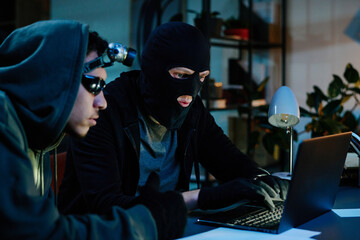 Young adult man wearing hoodie and man in balaclava sitting at desk using laptop, both focusing...