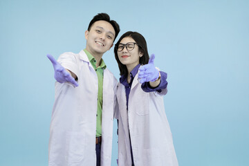 Smiling Asian science professionals giving a thumbs-up and an open-hand welcome gesture, symbolizing approval in blue studio background