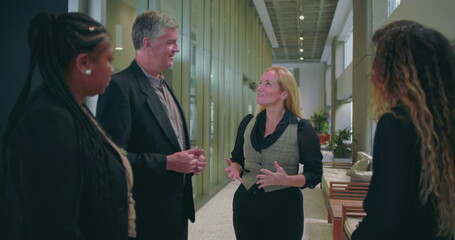 Confident female team member leading conversation with colleagues in modern office hallway, diverse group engaged in professional workplace discussion