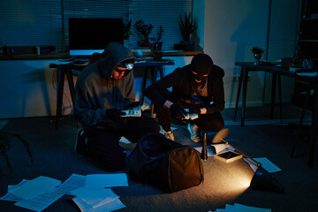 Two young adult men wearing masks and gloves, kneeling on office floor stealing cash from bag,...