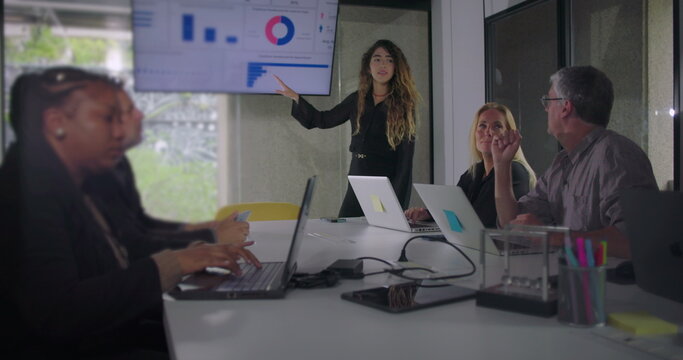 Confident woman stands in front of data screen as business team listens attentively during office meeting with laptops on table and focus on digital charts