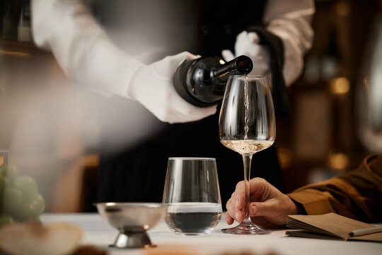 Caucasian man holding wine glass while sommelier in uniform pouring white wine during wine tasting session, closeup of hands and glassware on table, focus on tasting process