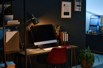 Dimly lit sht of modern office during night. Computer placed on wooden table, books and documents on shelves 