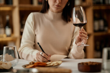 Young adult Caucasian woman holding wine glass and writing tasting notes at table with snacks, focusing on evaluating wine during tasting session, background showing wine shelves