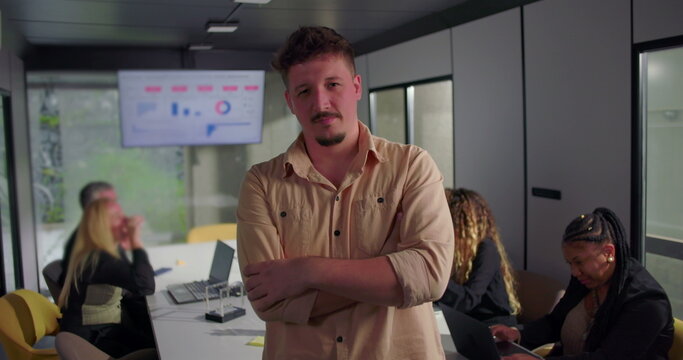 Confident young businessman with arms crossed in office, serious expression, diverse team focused on laptops in background near digital display
