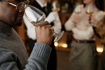 Black man smelling wine in glass during tasting session with two Caucasian young adults holding wine glasses in background, group participating in wine evaluation event