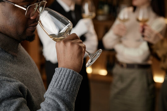 Black man smelling wine in glass during tasting session with two Caucasian young adults holding wine glasses in background, group participating in wine evaluation event