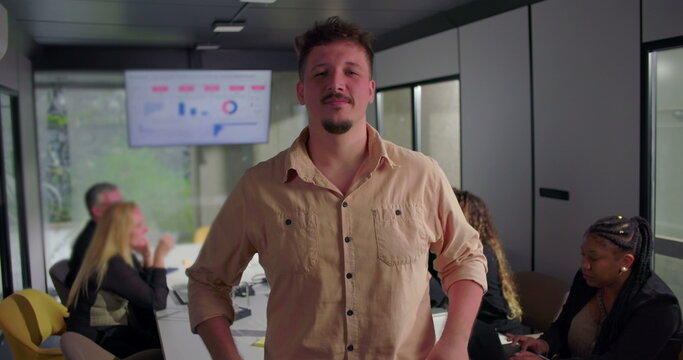 Confident young businessman with arms crossed in office, serious expression, diverse team focused on laptops in background near digital display