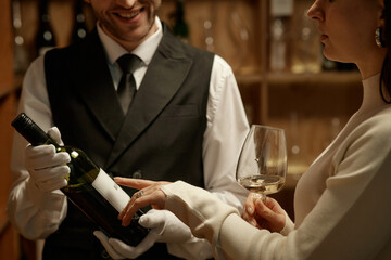 Caucasian young adult man wearing formal vest presenting wine bottle to Caucasian young adult woman holding wine glass, both engaging in wine tasting in cellar setting