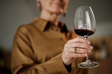 Middle aged Caucasian woman holding wine glass, examining red wine closely during tasting, hand with ring visible in foreground, blurred face and upper body in background