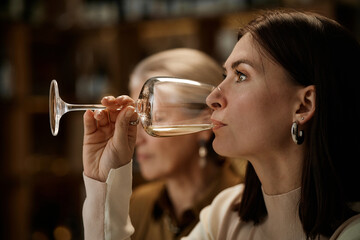 Young adult Caucasian woman tasting wine in profile with middle aged Caucasian woman in background participating in wine testing session, both focusing on evaluating flavor and aroma