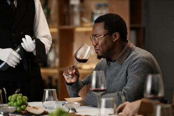 Black middle aged man sitting at table holding wine glass near face evaluating aroma during wine tasting session with server standing nearby, holding bottle and wearing gloves