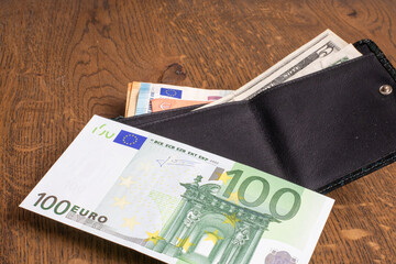 Black leather wallet with 100 Euro and US Dollar bills on wooden surface. Finance, currency, personal budget. Real photo