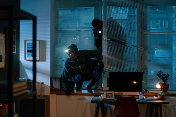 Two young adult men wearing hooded sweatshirts and headlamps, breaking into office through window...