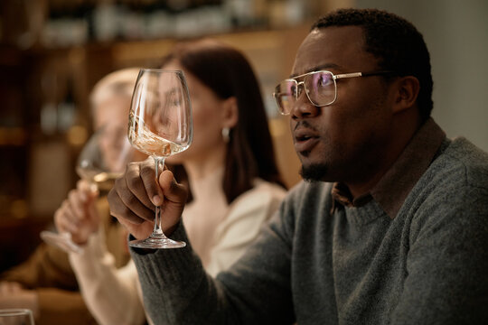 Middle aged Black man holding wine glass and examining color while sitting beside Caucasian woman and senior Caucasian woman, group participating in wine tasting session indoors
