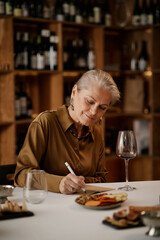 Senior Caucasian woman sitting at table tasting wine and writing notes, holding pen in hand with glass of red wine and plate of snacks in front, wooden wine shelves in background