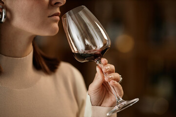 Caucasian young adult woman holding wine glass near face, smelling red wine during tasting, showing partial profile with focus on hand and glass, blurred background