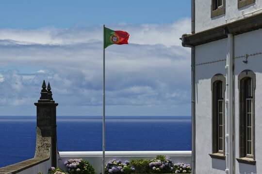 Portuguese flag flying next to a white building with a view of the blue sea and blooming flowers, Farol da Ferraria lighthouse, Sao Miguel Island, Azores, Portugal