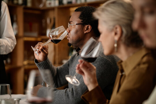Middle aged Black man and middle aged Caucasian woman sitting at table tasting red wine, holding glasses near faces, focusing on aroma, participating in wine tasting event