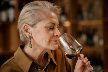 Naklejka premium Senior Caucasian woman holding wine glass, smelling red wine during tasting session, eyes closed, short gray hair, standing in front of blurred wine bottles on shelves