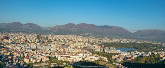 Naklejka premium A warm, sweeping panorama of Tirana's expansive cityscape and the Artificial Lake, set against the majestic Dajti Mountain range under the late afternoon light.