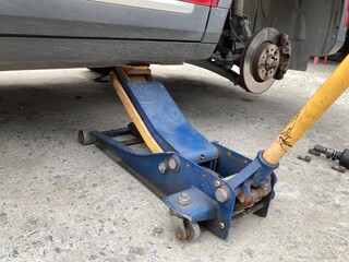 Low-angle view of a red car lifted by a hydraulic jack on a concrete floor. The wheel is removed, showing the brake disc and caliper assembly during a DIY repair or maintenance process.