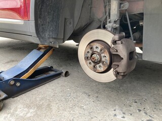 Low-angle view of a red car lifted by a hydraulic jack on a concrete floor. The wheel is removed, showing the brake disc and caliper assembly during a DIY repair or maintenance process.