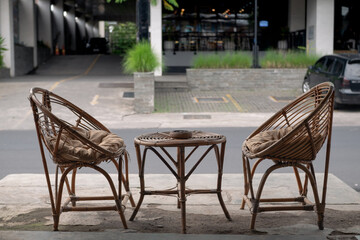 Modern outdoor rattan furniture set with two single chairs featuring beige cushions and one round table, background view with natural light and warm tones in a hotel parking.