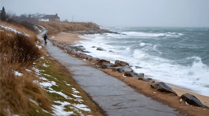 Solitary dog walker on a windswept winter coastal path with dramatic waves and scattered snow