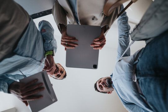 A diverse group of coworkers gathers in the office, looking up at a tablet as they discuss ideas and plan. Collaboration, technology, and shared goals define this workplace moment.