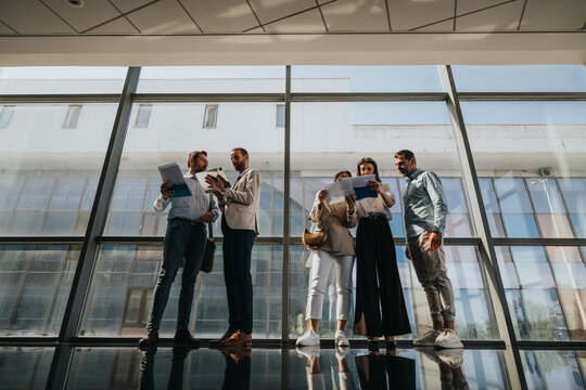 A team of colleagues stands in a bright glass corridor, reviewing papers and files. They share ideas, collaborate, and plan projects in a contemporary corporate setting.