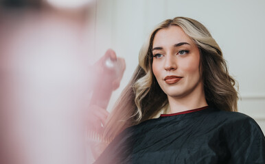 Fototapeta premium A client sits in a salon chair as a stylist styles her long hair, using a spray bottle. The calm, professional atmosphere highlights beauty, fashion, and grooming in a modern studio.