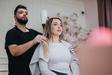 A male stylist uses a blow dryer on a seated female client in a contemporary salon. Soft lighting and floral decor create a calm, professional atmosphere as hair is dried and styled.