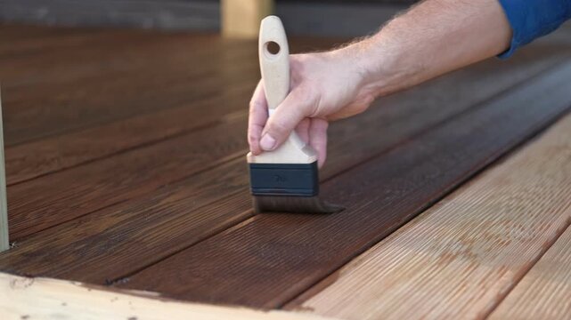 Brown oil for protection of wood by staining. Painting a larch for protecting and pests. Woodworker applying finish to terrace deck board. Repairman varnishing with brush wooden plank floor surface.