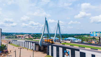 Aerial view of a yellow truck traversing a modern bridge with striking black and white patterns against a backdrop of lush greenery, Amawbia, Anambra, Nigeria.