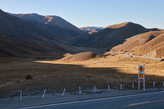Mackenzie Basin, New Zealand - May 23, 2025: Dry, tussock-covered rolling hills and mountains on the road to Mount Cook. Sunlight and shadow emphasize the dramatic, vast landscape, with a winding road