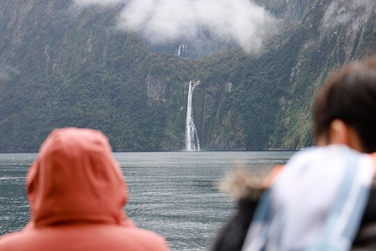 Milford Sound, New Zealand - May 20, 2025: A tall waterfall plunges down the sheer, mossy cliffs. Blurred tourists in the foreground view the dramatic landscape from a cruise boat.