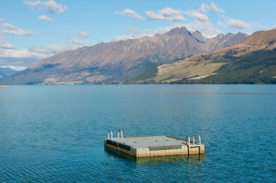 Glenorchy Pier, New Zealand - May 18, 2025: A floating dock sits in Lake Wakatipu with the autumn-colored forest and snow-capped mountains of Glenorchy visible in the distance under a blue sky.