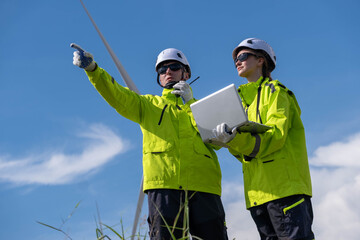 Engineers examine wind turbine site while analyzing data during bright sunny day with clear blue sky