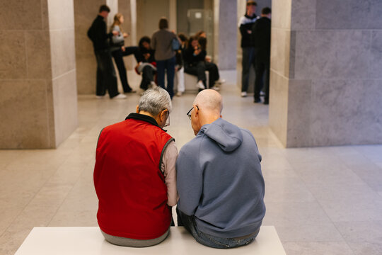 Two older people, a couple, sit on a bench in a museum.