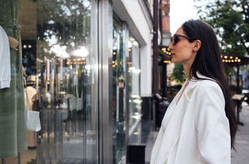 Stylish woman with long dark hair, wearing sunglasses and a white blazer, gazes thoughtfully at a boutique window display, showcasing elegant fashion items and accessories