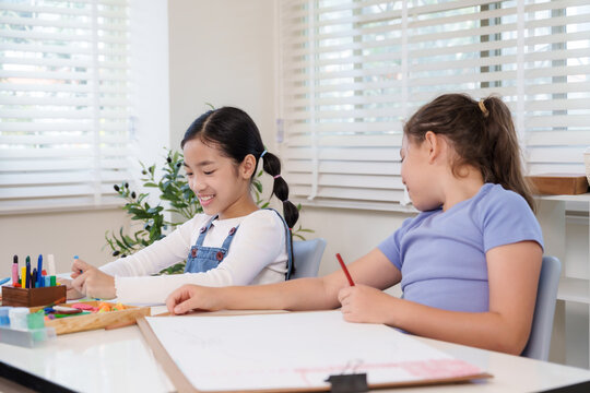 Asian and caucasian preteen classmates together at classroom desk collaborating on art worksheet drawing during elementary lesson teamwork creativity learning concentration diligence achievement