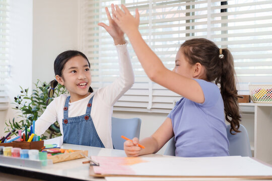Asian girl and caucasian girl elementary students smiling giving high five with excitement during classroom art project representing joy friendship teamwork childhood positive creative collaboration