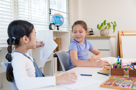 Asian girl preteen showing drawing paper smiling caucasian friend classroom desk during elementary art education teamwork creativity friendship playful childhood learning activity - Powered by Adobe