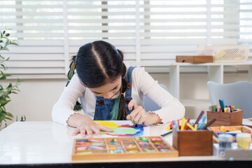 Asian preteen girl bending over paper coloring bright shapes with pastel during classroom art lesson surrounded by school supplies elementary creativity concentration diligence learning achievement